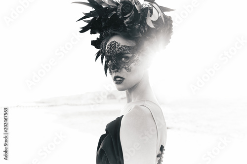 Black and white dramatic portrait of beautiful young woman in dress with black flowers and with black lace mask on face and floral crown on head. Sea background