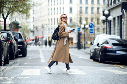 Pretty young woman crossing the street while holding the smartphone and looking sideways.