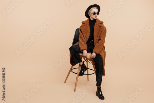 Serious young hipster woman with blonde short hair wearing a coat, hat and sunglasses posing over beige background.