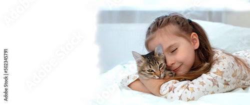 Banner. Portrait. Close-up. The child is resting on the bed with his kitten. A little girl in pajamas on a sunny morning with tenderness and love hugs a gray cat and smiles	
