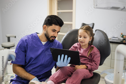 Medicine, pediatric dentistry and oral care concept. Confident male smiling professional dentist showing tablet pc computer to happy kid patient, caucasian preschool girl at modern dental clinic.