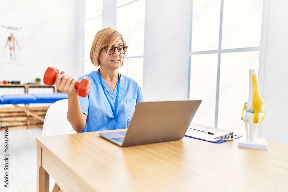 Middle Age Blonde Woman Wearing Physio Therapy Uniform Doing Tele Rehab Using Dumbbells At