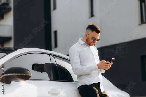 Portrait of handsome young businessman in black suit and tie outdoors near modern car in the city.