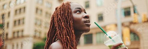 Panorama of beautiful woman with African braids dress wearing top walks down the street with cold drink in her hands. Stylish girl enjoys drinking fresh cocktail drink in plastic cup with straw