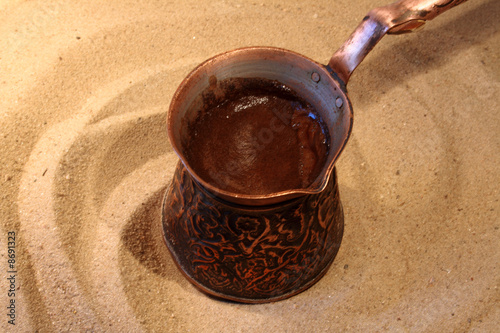 Naklejka dekoracyjna Black turkish coffee in brazen cezve standing on hot sand