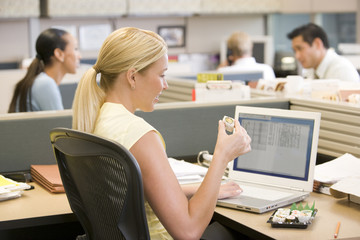 Wall Mural - Businesswoman in cubicle using laptop eating sushi