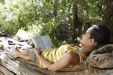 teenage girl (16-17 years) lying on tree trunk in forest reading book