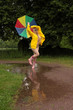 Eine Frau tanzt im Regen mit buntem Schirm und gelber Regenjacke über dem nackten Körper