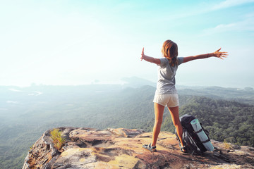 Poster - Young woman standing with raised hands with a backpack on the cliff's edge and looking into a wide valley