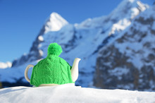 Tea Pot In The Knitted Cap On The Snow Against Mountain Peak