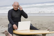 © REB Images/Blend Images - Senior Hispanic man sitting on surfboard using laptop and talking on cell phone