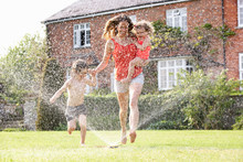 Mother And Two Children Running Through Garden Sprinkler