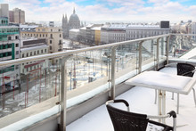 Table And Chairs On Balcony In Snow At Winter Day. View Of City.