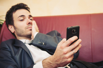 Portrait of tired young businessman typing on cellphone on sofa.