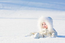 Baby In White Suit Sitting In Snow Field On Sunny Winter Day