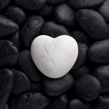 White Heart Rock On Black Rocks Background Or Texture. White Big Heart Pebble, Stone, Rock Laid On Pile Of Black Nature Pebbles, Stones, Rocks With Text Space. Heart Shape Of Pebble On Small Peddles.