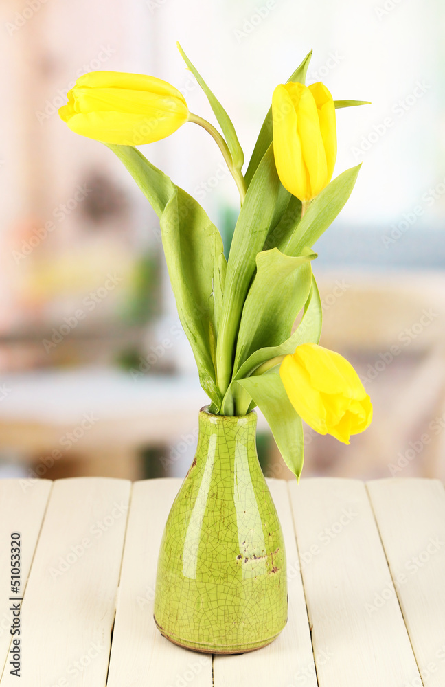 Yellow tulips in vase on wooden table on room background
