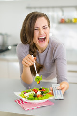 Wall Mural - Laughing young woman eating salad and watching tv in kitchen