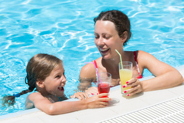 Mother and daughter swimming in pool with drink in hand