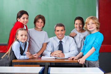 Confident Male Teacher With Schoolchildren  At Desk