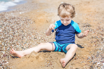 Wall Mural - Little toddler boy playing with sand and stones on the beach