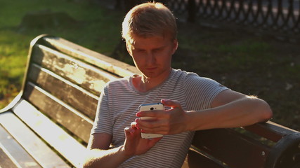 young blond adult man writing sms message in park bench