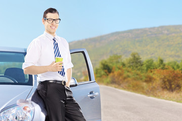 Canvas Print - Smiling young man on his automobile drinking coffee on a road