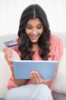 © lightwavemedia - Excited cute brunette sitting on couch holding credit card and t