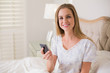 © lightwavemedia - Natural smiling woman sitting in bed holding smartphone