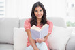 © lightwavemedia - Smiling cute brunette sitting on couch reading a book