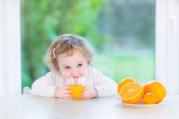 Cute toddler girl drinking orange juice sitting at a white table