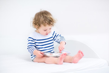 Cute toddler girl reading a book sitting in a white bedroom