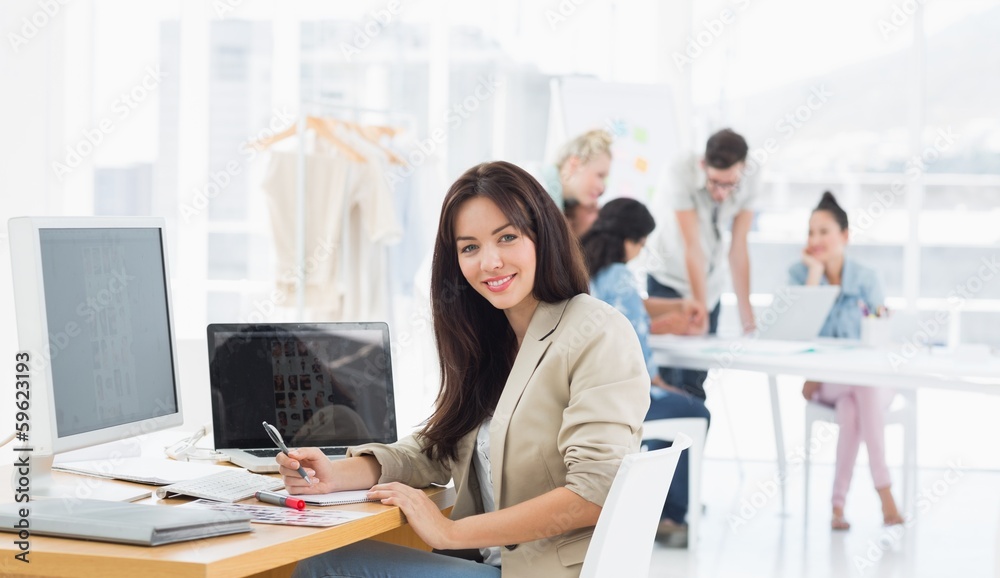 Casual woman at desk with colleagues behind in office - obrazy, fototapety, plakaty 
