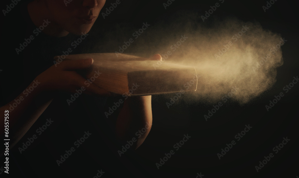 A stock photo of a person in a casual outfit blowing dust off the cover of an old, leather-bound book.
