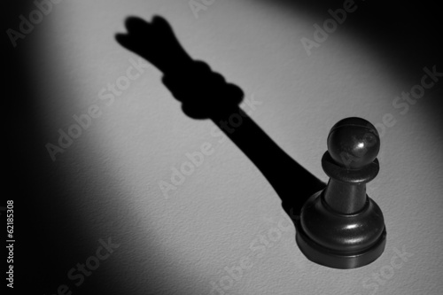 Fotografie, Obraz Chess pawn standing in a spotlight that make a shadow of queen a