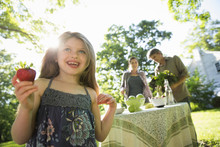 On The Farm. Children And Adults Together. A Young Girl Holding A Large Fresh Organically Produced Strawberry Fruit. Two Adults Beside A Round Table. 