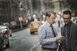 © Mint Images - Two men standing together looking at a cell phone display on a busy street at dusk.