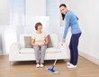 Caretaker Cleaning Floor While Woman Sitting On Sofa