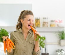 Happy Young Housewife Eating Carrot In Kitchen