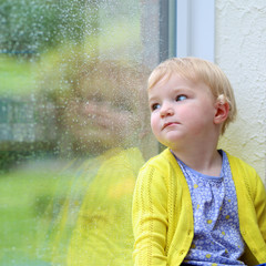 Wall Mural - Cute toddler girl sitting on the window looking outside at rain