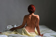 Back view of a naked woman sitting in bed covered with sheets