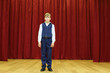 © Pavel Losevsky - Serious boy in suit with vest stands on stage with red curtains.