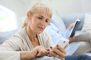 Wall Mural - Senior woman using smartphone, husband reading book