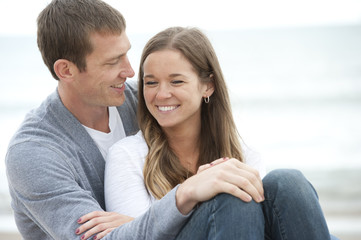 Wall Mural - A young happy caucasian couple sitting on the sand at the beach on a sunny day.