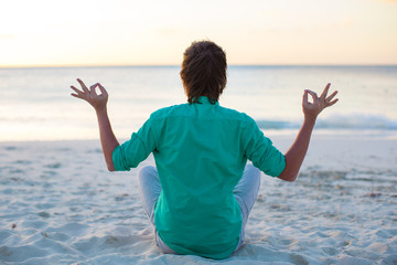 Wall Mural - Young man sitting in lotus position on white sand beach