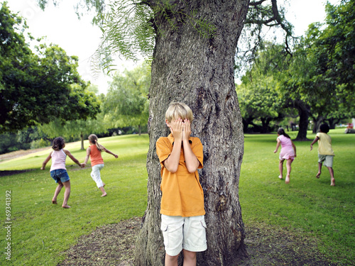 Fotomural Boy (7-9) with hands covering eyes playing hide and seek in park, hiding from fr