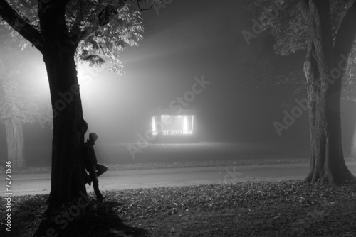 Stampe Man leaning agianst a tree in a foggy park at night.
