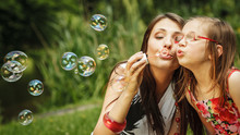 Mother And Little Girl Blowing Soap Bubbles In Park.