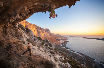 Rock climber climbing on roof in cave, his partner belaying