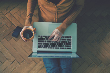 Laptop and coffee cup in girl’s hands sitting on a wooden floor